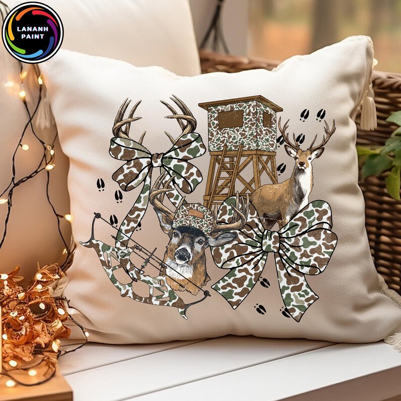 May include: Beige throw pillow with a hunting theme. The pillow features a deer, a hunting blind, a bow and arrow, and camouflage bow accents. The design includes deer tracks and the text "LANANH PAINT" in a circular logo.