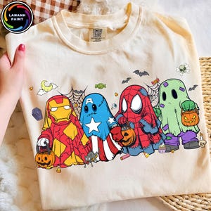 May include: A cream-colored t-shirt featuring a graphic of four characters dressed in Halloween ghost costumes resembling Iron Man, Captain America, Spider-Man, and a classic ghost. They carry pumpkin buckets amidst spiderwebs and candy.