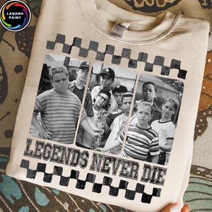 May include: Beige sweatshirt featuring a black and white image of a group of boys. The image is framed with a black and white checkered pattern. The text "LEGENDS NEVER DIE" is printed in black below the image.