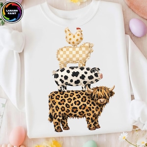 Op de afbeelding: Witte sweatshirt met een ontwerp van gestapelde boerderijdieren: een kip, een schaap, een varken en een koe. De koe heeft een luipaardprint, het varken heeft zwarte vlekken, het schaap heeft een geruit patroon en de kip heeft stippen. De sweatshirt heeft lange mouwen.