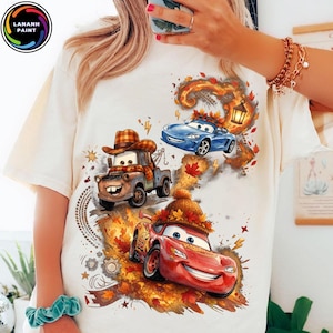 Puede incluir: Una camiseta de color crema con un estampado gráfico de tres coches animados. Una grúa marrón lleva un sombrero de cuadros, un coche deportivo azul está rodeado de hojas de otoño y un coche de carreras rojo lleva un gorro de punto entre hojas.