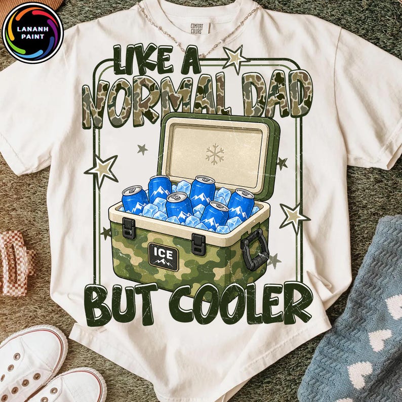 K&ouml;nnte beinhalten: Wei&szlig;es T-Shirt mit einem Grafikdesign, das einen Camouflage-K&uuml;hler mit Eis und blauen Dosen zeigt. Der Text lautet "LIKE A NORMAL DAD BUT COOLER" in gr&uuml;nen und braunen Buchstaben. Das Shirt hat einen Rundhalsausschnitt und kurze &Auml;rmel.