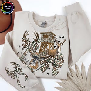May include: A beige sweatshirt featuring a deer-themed design. The graphic includes deer, a hunting blind, camouflage bows, and a bow and arrow. The sweatshirt has long sleeves and a crew neck, with the brand name "LANANH PAINT" in a circular logo.
