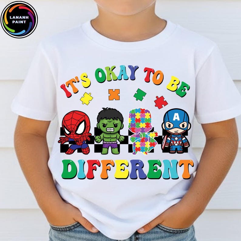 May include: White t-shirt with the text "It's Okay to Be Different" in rainbow colors. The shirt features cartoon illustrations of Spider-Man, Hulk, a puzzle piece figure, and Captain America. The shirt is for children.