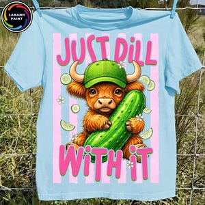 May include: Light blue t-shirt with a graphic of a Highland cow wearing a green cap and holding a large cucumber. The text "JUST DILL WITH IT" is in pink. White flowers and cucumber slices are also featured.
