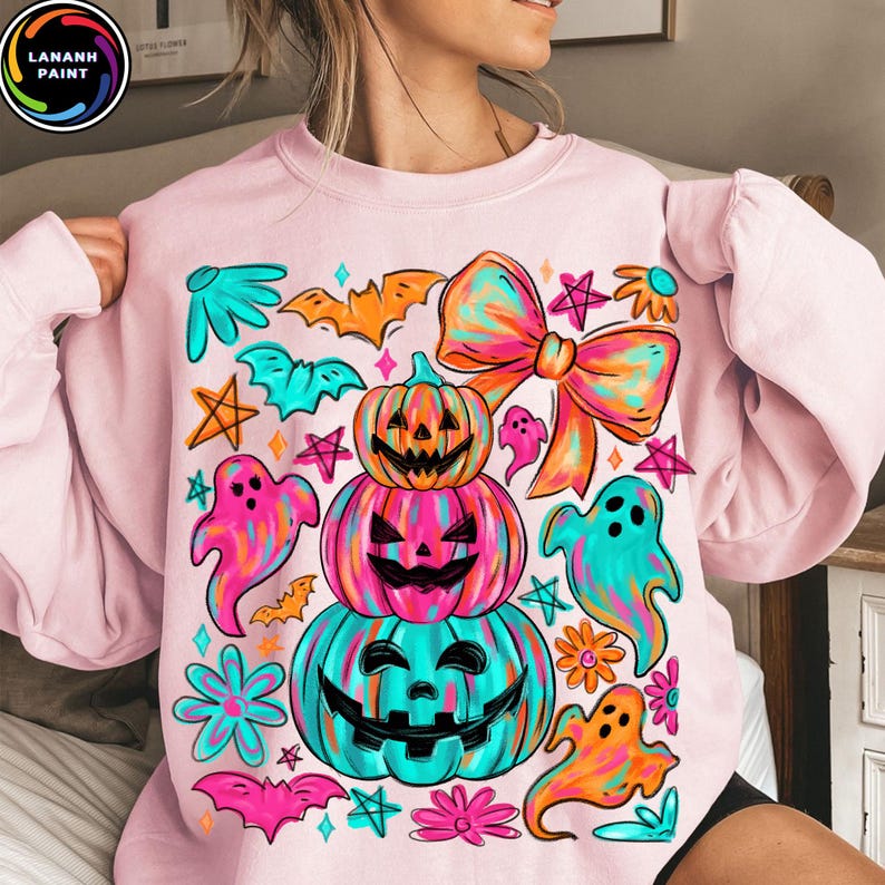 May include: Light pink sweatshirt with a colorful Halloween-themed graphic. The design features stacked jack-o'-lanterns, ghosts, bats, stars, flowers, and a large bow. The graphic is in shades of pink, orange, teal, and purple.