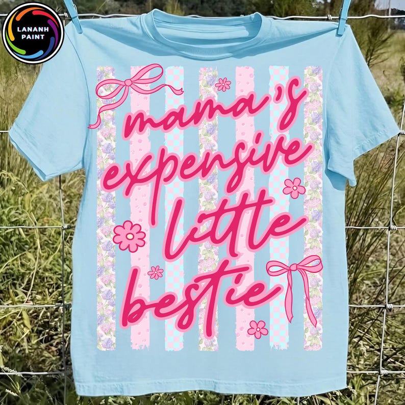 Puede incluir: Camiseta azul claro con el texto "mama's expensive little bestie" en escritura rosa. La camiseta presenta rayas verticales, lazos y detalles florales. La camiseta est&aacute; colgada en un tendedero.