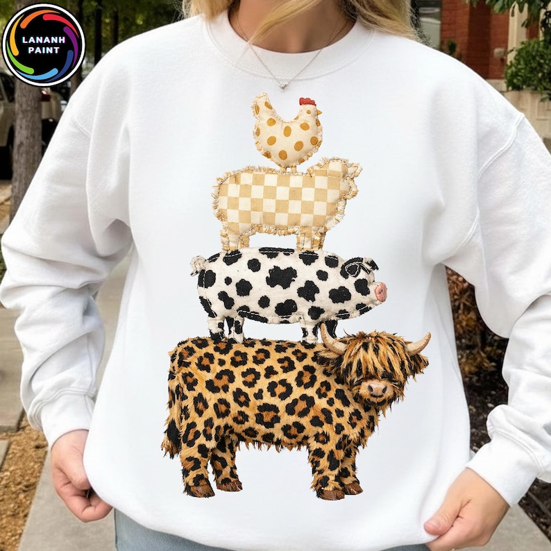 Op de afbeelding: Witte sweatshirt met een ontwerp van boerderijdieren op elkaar gestapeld: een Schotse hooglander met een luipaardprint, een varken met zwarte vlekken, een schaap met een geruit patroon en een kip met stippen. Het sweatshirt heeft de tekst "LANANH PAINT".