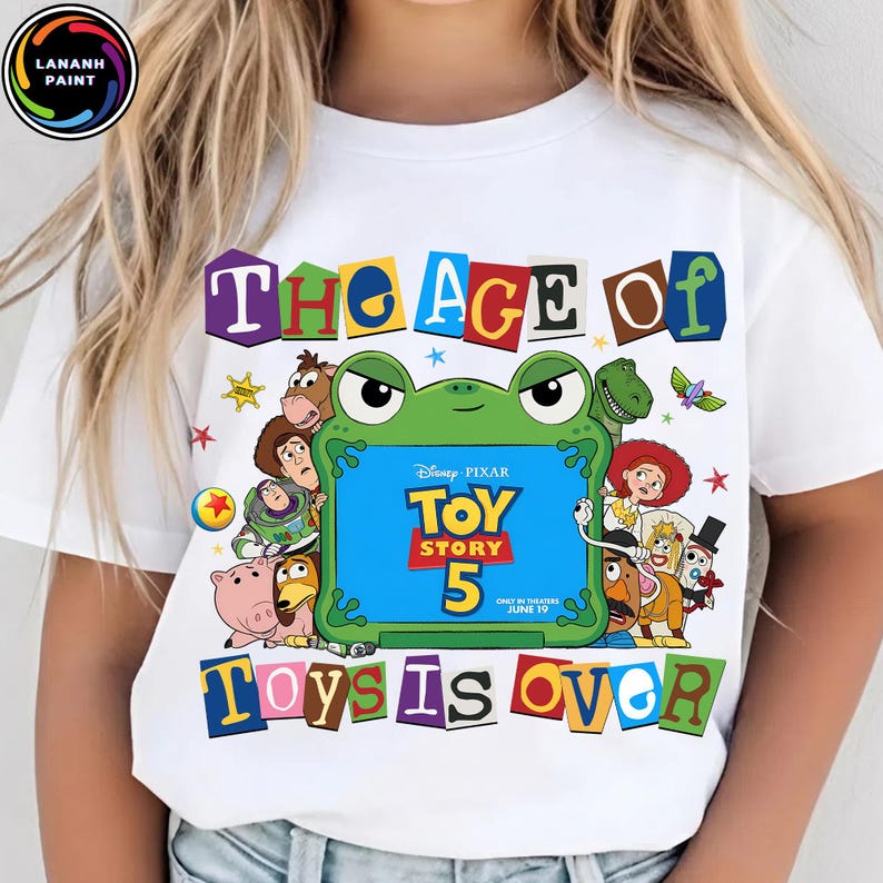 Puede incluir: Una camiseta blanca con un estampado gr&aacute;fico que incluye el texto THE AGE OF TOY STORY 5 TOYS IS OVER. El dise&ntilde;o presenta un marco central con forma de rana verde que rodea el logotipo de Toy Story 5, junto a varios personajes animados.