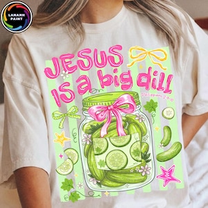 May include: A cream-colored t-shirt featuring a graphic of a glass jar filled with green pickles. The text reads "JESUS is a big dill" and "Philippians 2:9-10" in pink. The design includes a pink bow on the jar, yellow ribbons, and stars.