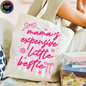 Puede incluir: Un bolso tote beige con el texto "mama's expensive little bestie" en escritura rosa. El dise&ntilde;o incluye lazos rosas, flores y rayas verticales en rosa y azul claro. El bolso tiene un asa blanca.