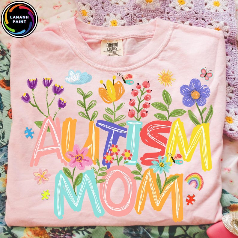 以下が含まれることがあります： 明るいピンク色のTシャツ。カラフルで気まぐれな文字で「AUTISM MOM？」と書かれています。デザインには花、蝶、虹、その他の装飾要素が含まれています。柔らかい素材で作られたシャツです。