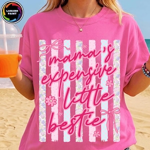 Puede incluir: Una camiseta rosa con la frase "mama's expensive little bestie" en cursiva. El dise&ntilde;o incluye rayas verticales, lazos y detalles florales. La camiseta es de cuello redondo.