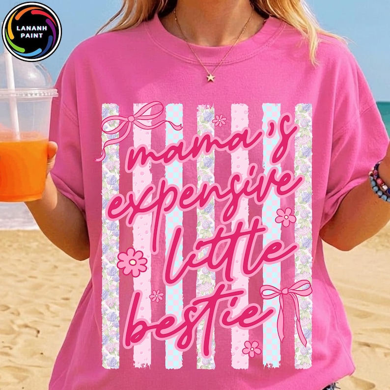 Puede incluir: Una camiseta rosa con la frase "mama's expensive little bestie" en cursiva. El dise&ntilde;o incluye rayas verticales, lazos y detalles florales. La camiseta es de cuello redondo.