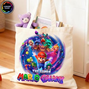 Puede incluir: Un bolso tote de color crema con un gr&aacute;fico colorido de Mario y otros personajes del juego Super Mario Galaxy. La bolsa tiene un oso de peluche morado y libros dentro. El texto "THE SUPER MARIO GALAXY" se muestra en una fuente vibrante y multicolor.