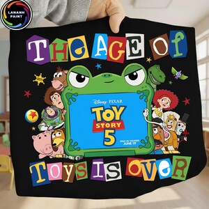 Puede incluir: Un art&iacute;culo de tela negra con un marco central en forma de rana verde que contiene el texto Disney Pixar Toy Story 5 Only in Theaters June 19. Alrededor del marco hay varios personajes animados y el texto The Age of Toys is Over en letras coloridas.
