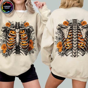 May include: A beige crewneck sweatshirt featuring a graphic print of a human ribcage and spine filled with orange jack-o'-lantern pumpkins, surrounded by detailed black and white floral illustrations and orange flowers on the front and back.
