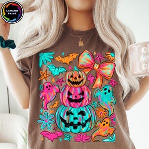 May include: Brown t-shirt with a colorful Halloween graphic. The design features stacked jack-o'-lanterns, ghosts, bats, and a large bow. The shirt has the text "LANANH PAINT" in a circle on the top left.