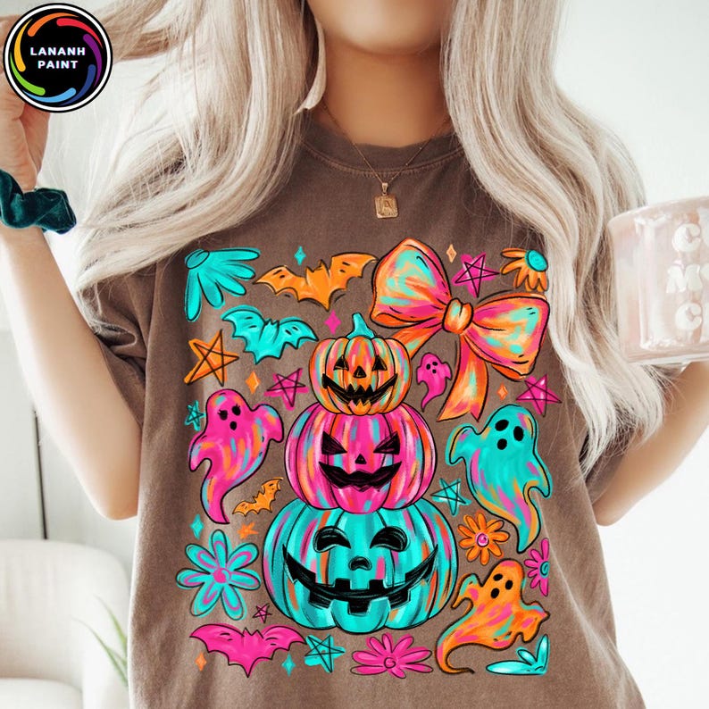 May include: Brown t-shirt with a colorful Halloween graphic. The design features stacked jack-o'-lanterns, ghosts, bats, and a large bow. The shirt has the text "LANANH PAINT" in a circle on the top left.