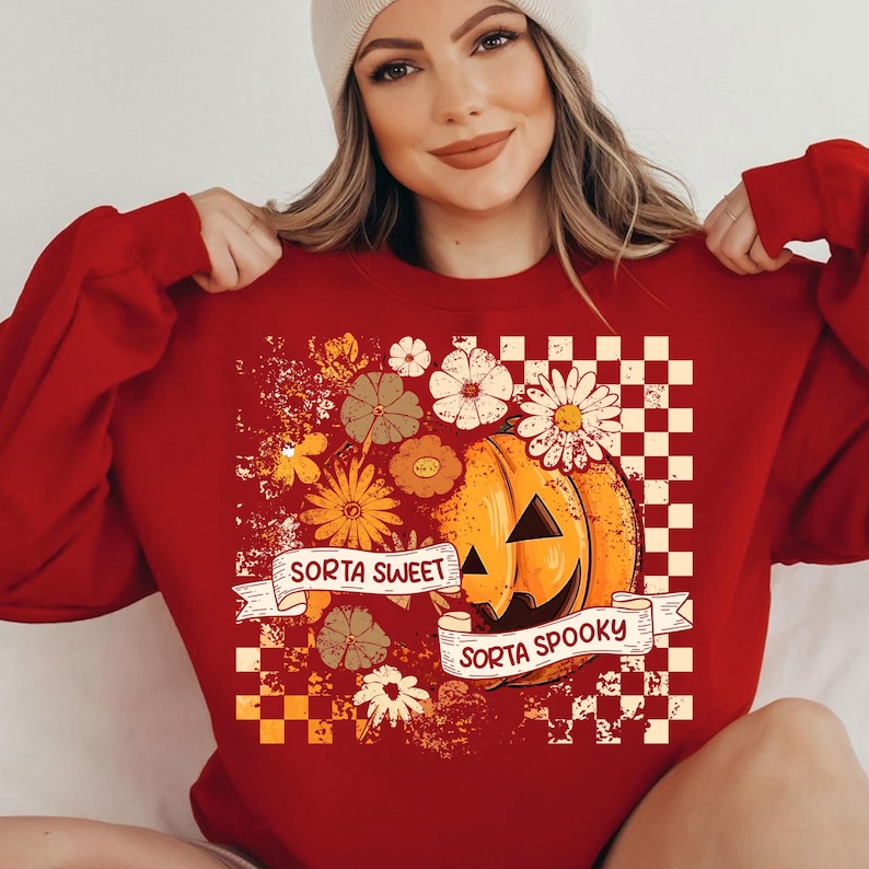 May include: A red sweatshirt featuring a pumpkin graphic with the text "Sorta Sweet Sorta Spooky." The design includes flowers and a checkered pattern. The pumpkin has a smiling face and is surrounded by floral elements.