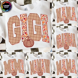May include: A white crewneck sweatshirt featuring the word GIGI in large block letters with a brown gingham and autumn floral pattern. A small orange pumpkin graphic is positioned below the text. The design includes a fall-themed aesthetic.