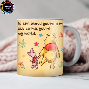 Op de afbeelding: Een keramische mok met Winnie de Poeh en Knorretje die elkaars hand vasthouden, met de tekst "To the world you're a mi but to me, you're my world." De mok heeft een gele achtergrond met bloemmotieven en een wit handvat.