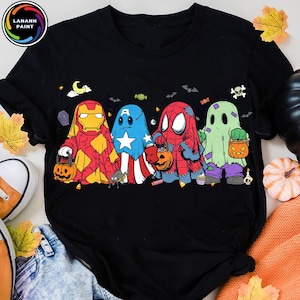 May include: A black short-sleeve t-shirt featuring a graphic of four characters dressed as ghosts for Halloween. The characters resemble Iron Man, Captain America, Spider-Man, and a green ghost, each holding a small orange jack-o'-lantern bucket.