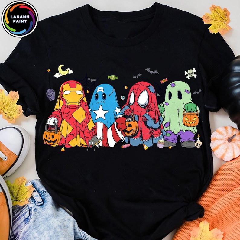 May include: A black short-sleeve t-shirt featuring a graphic of four characters dressed as ghosts for Halloween. The characters resemble Iron Man, Captain America, Spider-Man, and a green ghost, each holding a small orange jack-o'-lantern bucket.