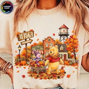 May include: A cream-colored t-shirt featuring a graphic of Winnie the Pooh and Piglet in an autumn scene. The design includes a wooden sign reading 47567 Petersburg, a water tower, houses, pumpkins, and fall foliage in shades of orange and red.