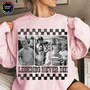 May include: A light pink sweatshirt featuring a black and white image of a group of boys. The image is framed with a black and gray checkered pattern. The text "LEGENDS NEVER DIE" is printed in white below the image.