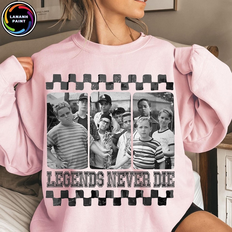 May include: A light pink sweatshirt featuring a black and white image of a group of boys. The image is framed with a black and gray checkered pattern. The text "LEGENDS NEVER DIE" is printed in white below the image.
