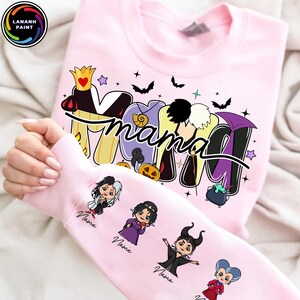 May include: A light pink sweatshirt with the word "MAMA" in a colorful, cartoonish font, with Halloween-themed accents. The sleeve features cartoon illustrations of Disney villains. The sweatshirt has a crew neck.