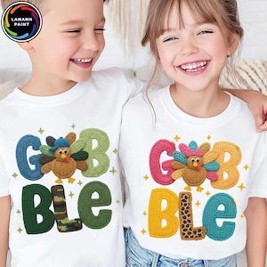 Könnte beinhalten: Zwei weiße Kinder-T-Shirts mit einem farbenfrohen "GOBBLE"-Design. Das Design zeigt einen gestrickten Truthahn und Buchstaben in Blau, Grün, Rosa, Gelb und Braun. Ideal für Thanksgiving.