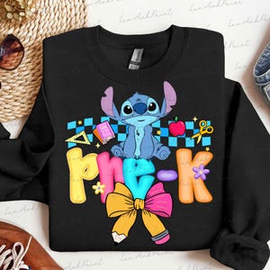 May include: A black crewneck sweatshirt featuring a graphic of the blue character Stitch sitting above the text "Pre-K". The design includes a blue checkered pattern, a red apple, a yellow ruler, a pink book, yellow scissors, and a large yellow bow.