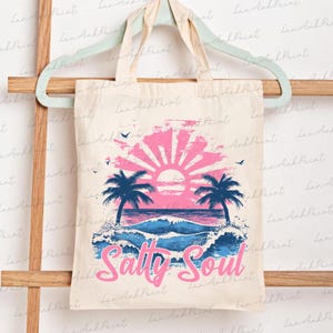 Pu&ograve; includere: Una borsa tote bianca con una grafica di tramonto rosa e blu e palme. Il testo "Salty Soul" &egrave; stampato in rosa sulla borsa.