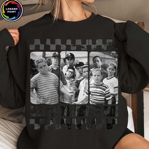 May include: Black sweatshirt featuring a black and white image of a group of boys. The image is framed by a checkered pattern. The text "The Sandlot" is printed at the bottom of the image. A circular logo is in the upper left corner.