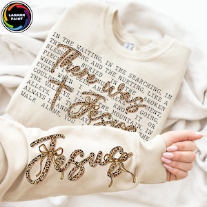 May include: A cream-coloured sweatshirt featuring the text "There is Jesus" in a leopard print design. The sleeve also displays the word "Jesus" in the same style. The sweatshirt has a crew neck and long sleeves.
