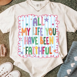 May include: Cream-colored t-shirt with the text "ALL MY LIFE YOU HAVE BEEN FAITHFUL" in colorful, playful lettering. The text is set against a light blue striped background with stars and a pink curly border. A casual, comfortable tee.
