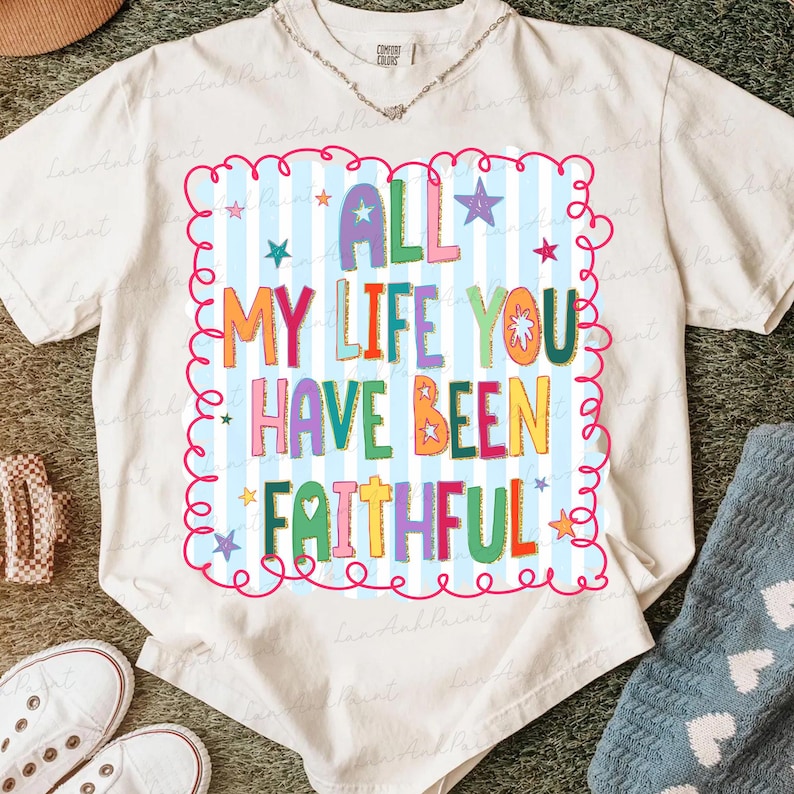 May include: Cream-colored t-shirt with the text "ALL MY LIFE YOU HAVE BEEN FAITHFUL" in colorful, playful lettering. The text is set against a light blue striped background with stars and a pink curly border. A casual, comfortable tee.
