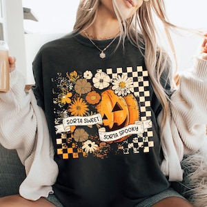 May include: Black sweatshirt with a pumpkin graphic, flowers, and the text "Sorta Sweet Sorta Spooky." The design includes a checkered pattern and a vintage aesthetic, perfect for Halloween.