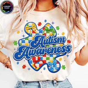 May include: Cream-colored t-shirt featuring a colorful puzzle piece design with the words "Autism Awareness" in blue script. The puzzle piece has a checkered pattern and is adorned with bows and stars.