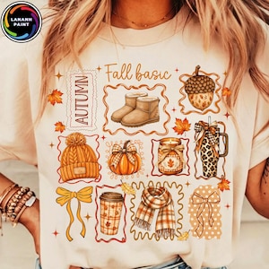 May include: A cream-coloured t-shirt featuring a graphic print with the text Fall basic and Autumn. The design includes illustrations of tan boots, an acorn, a knit beanie, a pumpkin, a candle, a leopard print tumbler, a coffee cup, and a scarf.