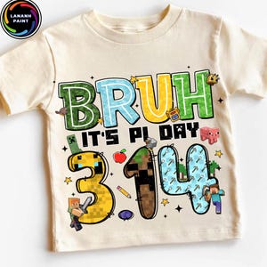 May include: Cream-colored t-shirt with a colorful graphic design. The design features the word "BRUH" in green, blue, and yellow, along with the phrase "IT'S PI DAY" and the numbers "3.14". Minecraft-style characters and icons are also included.