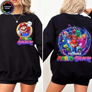 Puede incluir: Sudadera negra con Mario de Super Mario Galaxy. El dise&ntilde;o frontal muestra a Mario en un c&iacute;rculo dorado con el texto "The Super Mario Galaxy". El dise&ntilde;o trasero muestra a Mario y otros personajes en un dise&ntilde;o de galaxia colorido con el mismo texto.