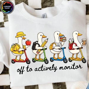 May include: White sweatshirt featuring a cartoon design of four ducks riding scooters. Each duck carries school supplies like an apple, pencil, books, and coffee. The text "off to actively monitor" is printed below the ducks.