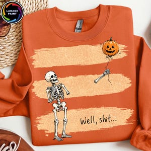 May include: An orange crewneck sweatshirt featuring a graphic of a skeleton looking at a detached skeletal arm holding a jack-o'-lantern balloon. The design includes three horizontal brushstroke textures with spiderweb patterns and the text Well, shit...