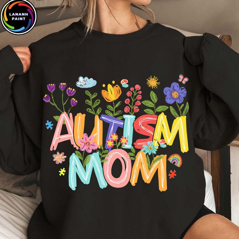 以下が含まれることがあります： カラフルで気まぐれな文字で「AUTISM MOM」と書かれた黒いスウェットシャツ。花柄と虹のグラフィックで囲まれています。デザインには、さまざまな花、雲、虹が含まれています。