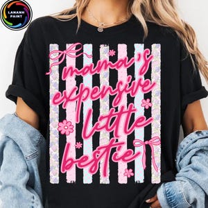 Puede incluir: Camiseta negra con la frase "Mama's Expensive Little Bestie" en rosa. El dise&ntilde;o incluye lazos rosas, flores y rayas verticales en rosa, blanco y azul claro.