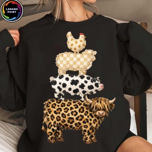 Op de afbeelding: Zwart sweatshirt met een stapelontwerp van boerderijdieren: een kip, een schaap, een koe en een Schotse Hooglander. De dieren zijn ontworpen met verschillende patronen, waaronder stippen, ruitjes en luipaardprint. Ronde hals.