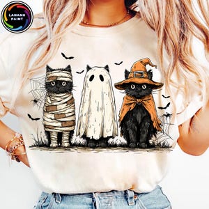 May include: Cream-colored t-shirt featuring three black cats dressed in Halloween costumes. One cat is wrapped in bandages, another is a ghost, and the third wears a witch hat and cape. Bats and spiderwebs complete the spooky design.
